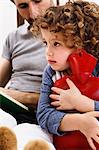 Girl hugging hot water bottle while father reads storybook in bed