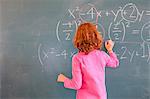 Rear view of primary schoolgirl answering equation on classroom blackboard