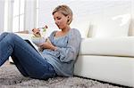 Mid adult woman sitting on rug looking at digital tablet and eating salad