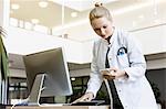 Doctor standing beside computer, holding open diary, looking at smartphone