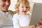 Female toddler sitting on father's knee using digital tablet