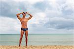 Muscular male swimmer standing on beach putting on swimming goggles