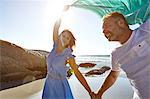 Mature couple holding hands on beach, woman holding sheer scarf in air