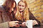 Two female friends, sitting outdoors, having coffee, looking at smartphone