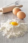 An egg yolk in a pile of flour with whole eggs and a rolling pin