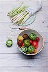 White and green asparagus on a wooden board, asparagus peelings, a peeler and a bowl of various tomatoes