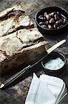 An arrangement of crusty bread, olives, sea salt, a knife and a fabric napkin