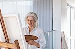 Smiling mature woman with palette painting at canvas on easel