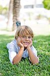 Cute blonde boy lying down on grass in park smiling at camera