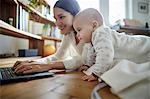 Baby daughter watching mother typing on laptop on floor