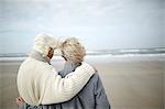 Pensive senior couple hugging and looking at ocean view on windy winter beach
