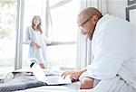 Smiling woman watching boyfriend in bathrobe reading laptop on bed