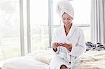 Smiling woman in bathrobe and hair wrapped in towel using digital tablet on bed