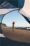 Man framed by camping tent, standing on beach and looking through binoculars at dusk, Olympic National Park, Washington, USA.