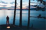 Man standing by campfire at dusk, San Juan Islands in the distance, Washington, USA.