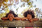 Portrait of two young sisters leaning against fence, covering eyes