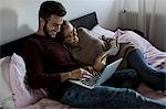 Young couple, relaxing on bed, young man using laptop, young woman reading book