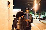 Couple walking in street at night looking at smartphone, Los Angeles, California, USA