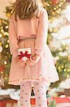 Girl in pink dress holding Christmas gift behind her back near Christmas tree