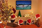 Family in Santa hats watching summer holiday video on TV in Christmas living room