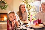 Portrait playful girl in paper crown blowing party favor at Christmas dinner table