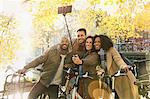 Smiling friends with bicycle taking selfie with selfie stick on urban bridge