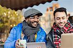 Smiling young men friends drinking coffee and using laptop and digital tablet at sidewalk cafe