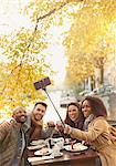 Smiling friends taking selfie with selfie stick at autumn sidewalk cafe