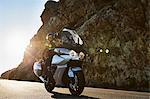 Couple riding motorcycle on sunny road along craggy cliff