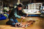 Man in a sailmaker's workshop kneeling on the floor, tying a rope on a sail.