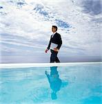 A man in a suit walking along a swimming pool, carrying a laptop.