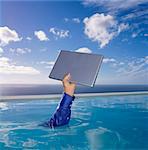 Human arm in blue shirt sleeve in swimming pool, holding aloft a laptop computer, sky and clouds.
