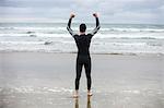 Rear view of athlete in wet suit standing with arms up on beach