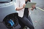 Mid section of woman using digital tablet while charging electric car on street