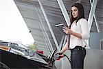 Beautiful woman using digital tablet while charging electric car at vehicle charging station