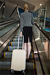 Rear view of businesswoman standing on escalator with luggage at airport