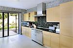 The interior spaces of a modern house on the cliffs overlooking the sea. Large glass panels and walls, and wooden shelves and room dividers. Kitchen area.