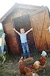 Little boy happily throwing seeds to the chickens in front of a henhouse