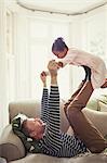 Multi-ethnic father playing, balancing daughter on legs overhead on sofa