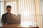 Male executive using laptop near window blinds in office