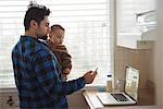 Father using mobile phone while holding his baby in kitchen at home