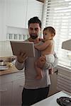 Father using digital tablet while holding his baby in kitchen