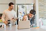 Male gay parents and baby son using laptop and digital tablet in kitchen