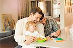 Male gay parents feeding baby son and using digital tablet