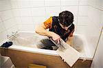 A builder, tiler sitting in a bathtub spreading adhesive on a tile.