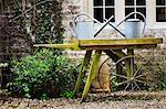 Old fashioned wooden wheel barrow and tin watering cans in the garden of an old house.