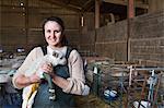 Smiling woman standing in a stable, holding a newborn lamb with bandaged front legs.