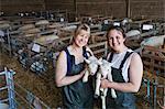 Two smiling women standing in a stable, holding newborn lambs.