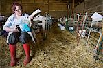 Young woman sitting in a barn, feeding a newborn lamb with milk from a bottle. Lamb dressed in a knitted blue jumper.