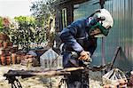 Man standing outdoors, wearing a face mask, working on a metal pitchfork with an angle grinder.